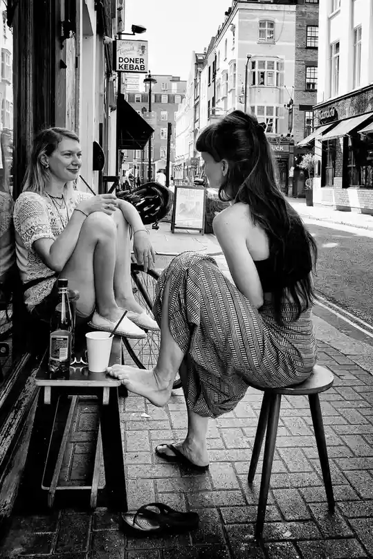 A black-and-white photo shows two women sitting on stools outside a cafe on a city street. One woman faces the camera with a…