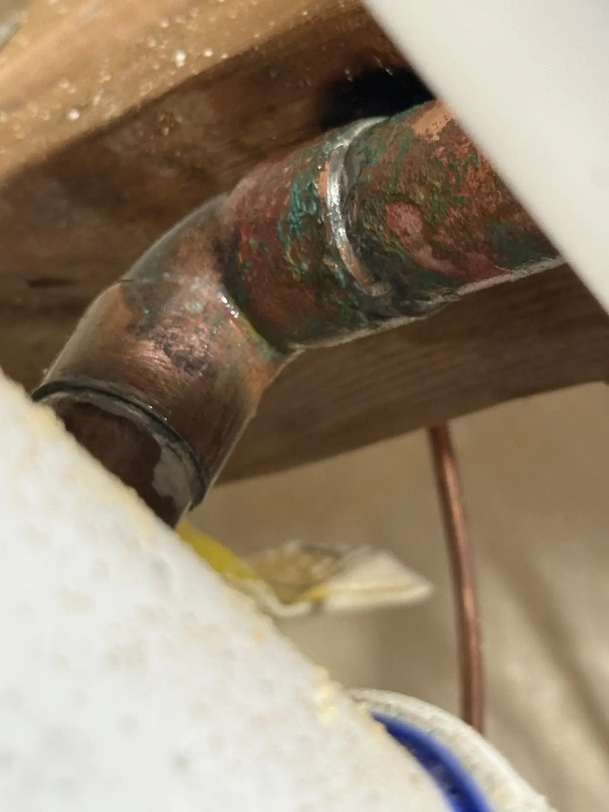 A close-up photo shows a corroded copper pipe joint under a sink, covered in green patina and water stains, with wooden…