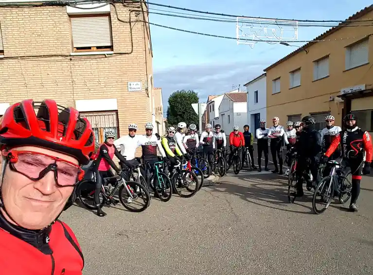 A cyclist in a red helmet and glasses takes a selfie in the foreground, smiling smugly, while behind him a group of cyclists…
