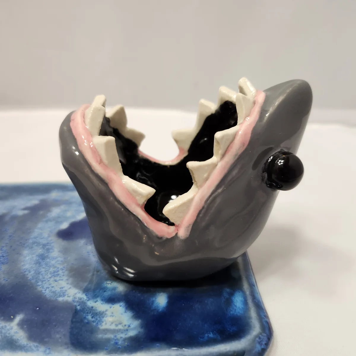 A ceramic figurine shaped like a shark's head with its mouth wide open, revealing sharp white teeth and pink gums, sits on a…