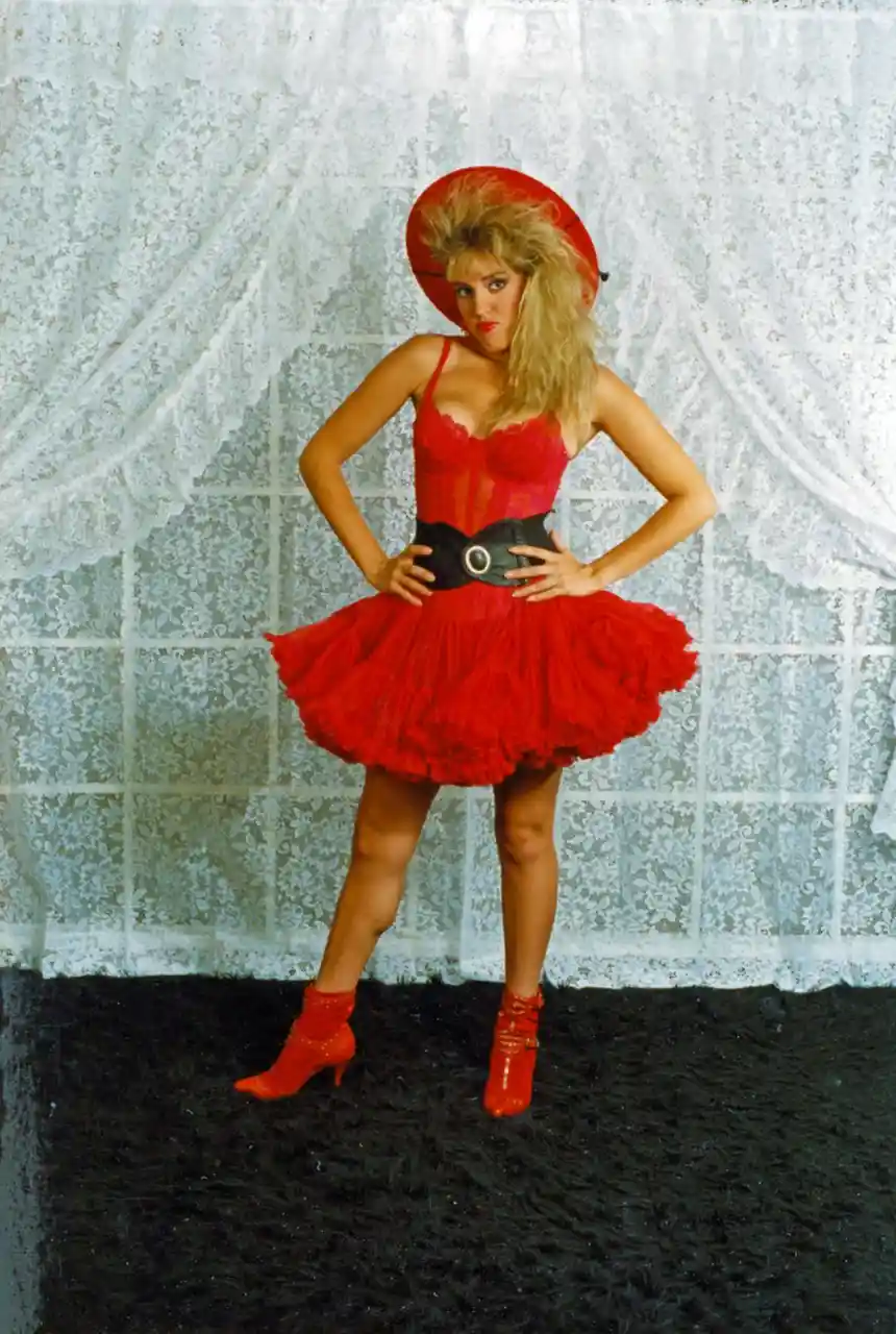 A woman with voluminous blonde hair poses confidently in a full red outfit including a corset, tutu skirt, and high-heeled…