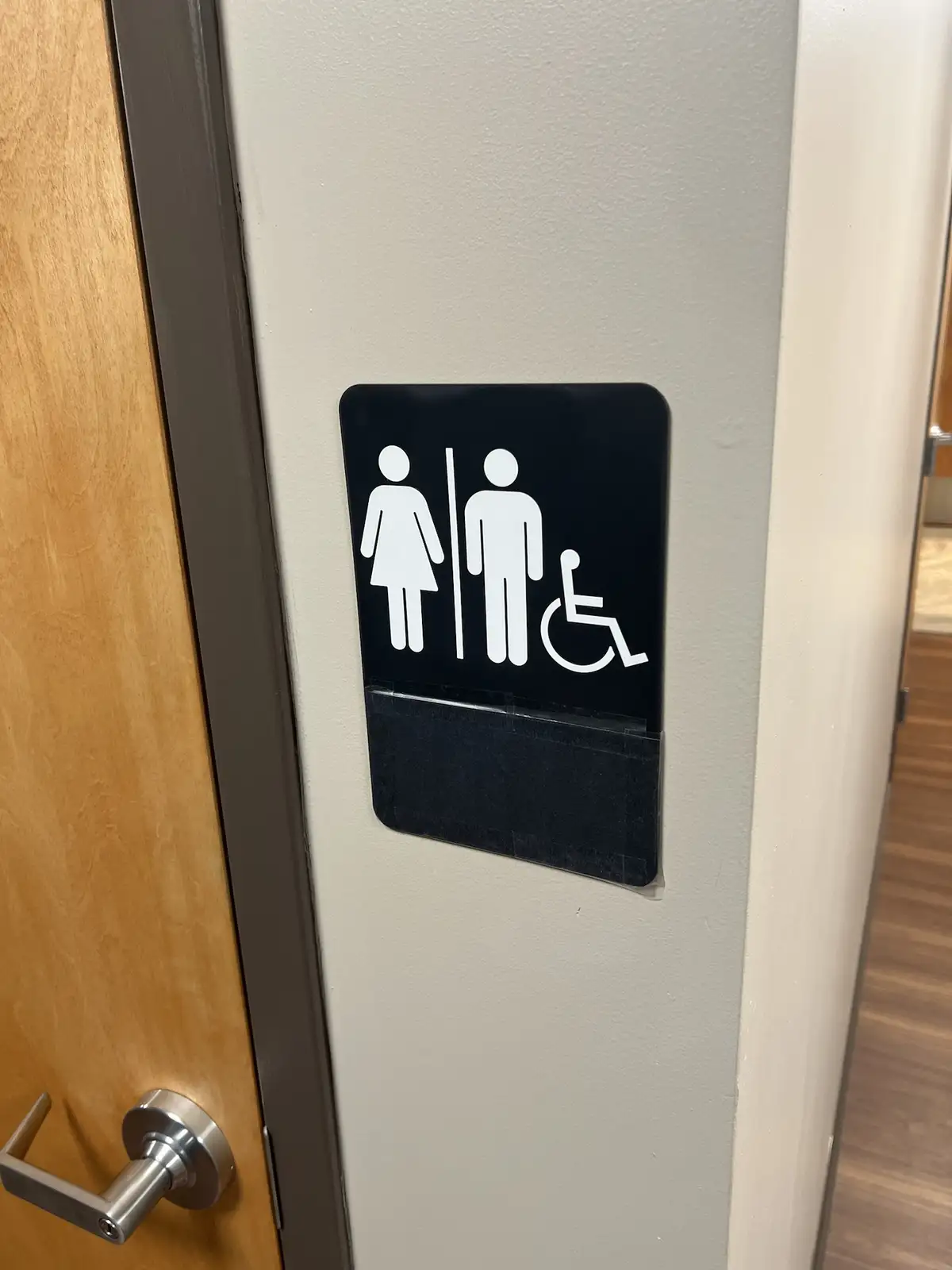 a close-up photo shows a black restroom sign on a beige wall next to a wooden door with a silver handle. The sign displays…