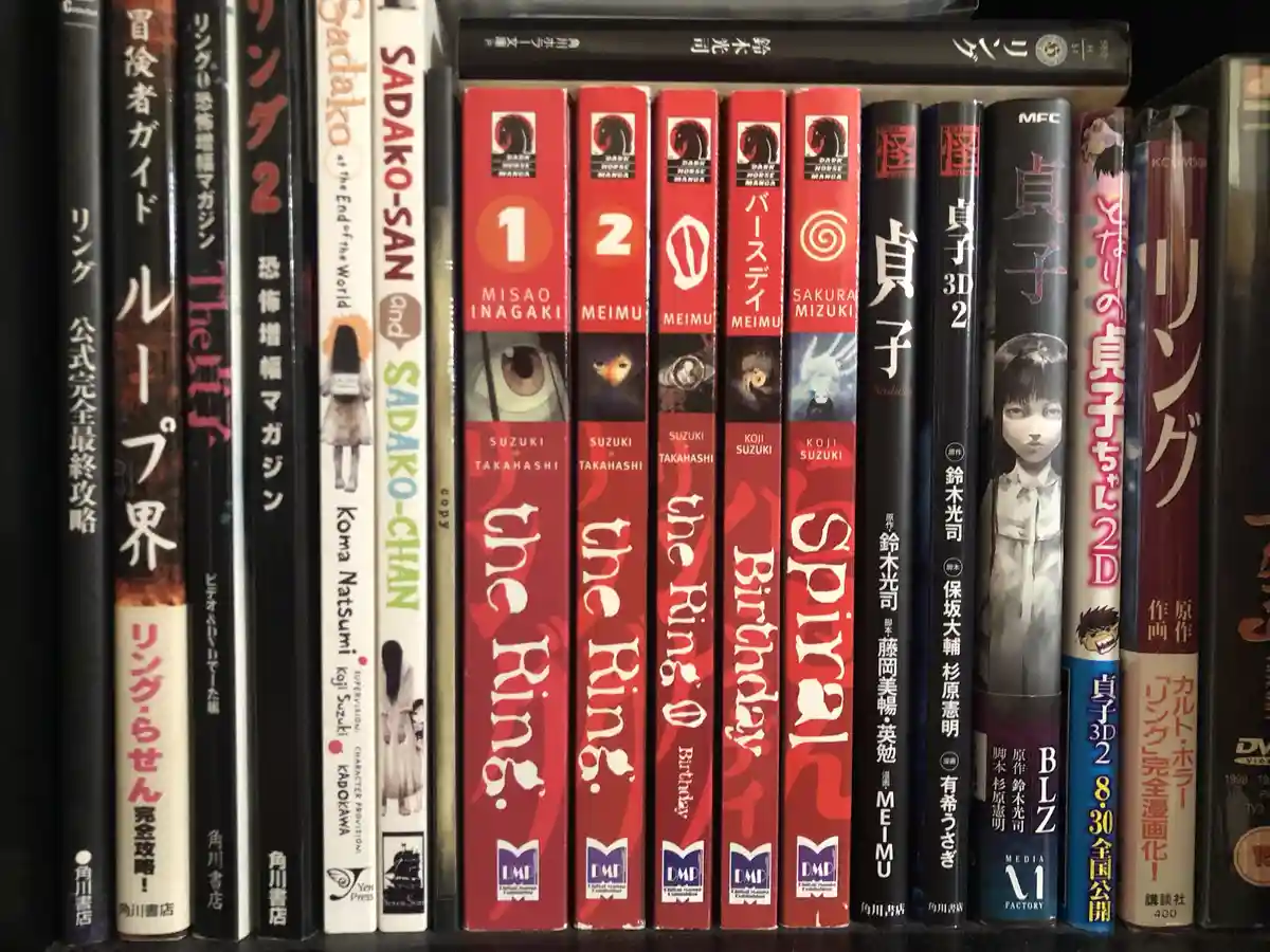 A close-up shot of a bookshelf filled with Japanese horror manga and media, prominently featuring multiple volumes of 'The…