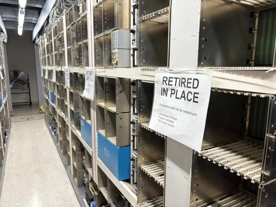 Rows of empty server racks in a data center, with a prominent white sign taped to one rack reading 'RETIRED IN PLACE' and…