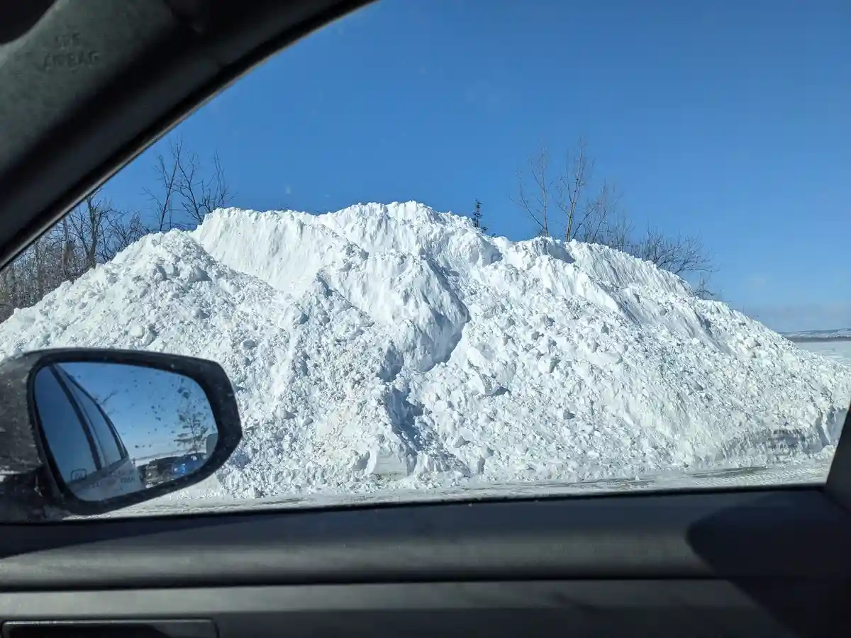 A photo taken from inside a car shows a massive, jagged pile of snow dominating the view through the window, with bare trees…