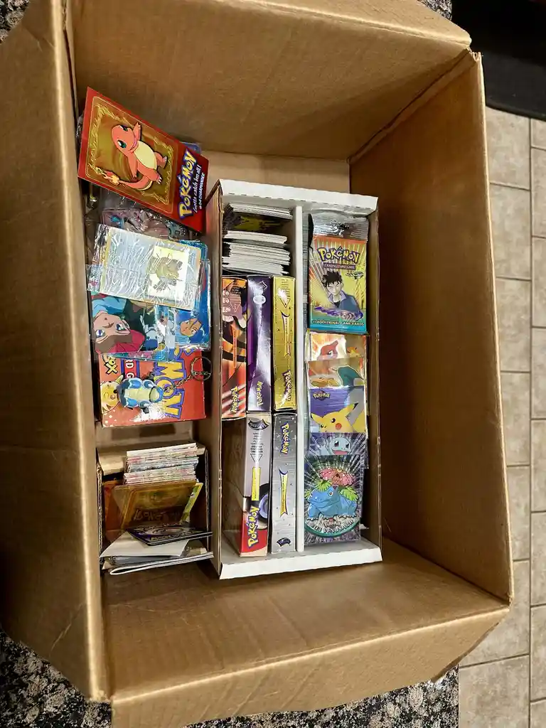 A large cardboard box is filled with an overwhelming amount of Pokémon trading cards, booster packs, and related…