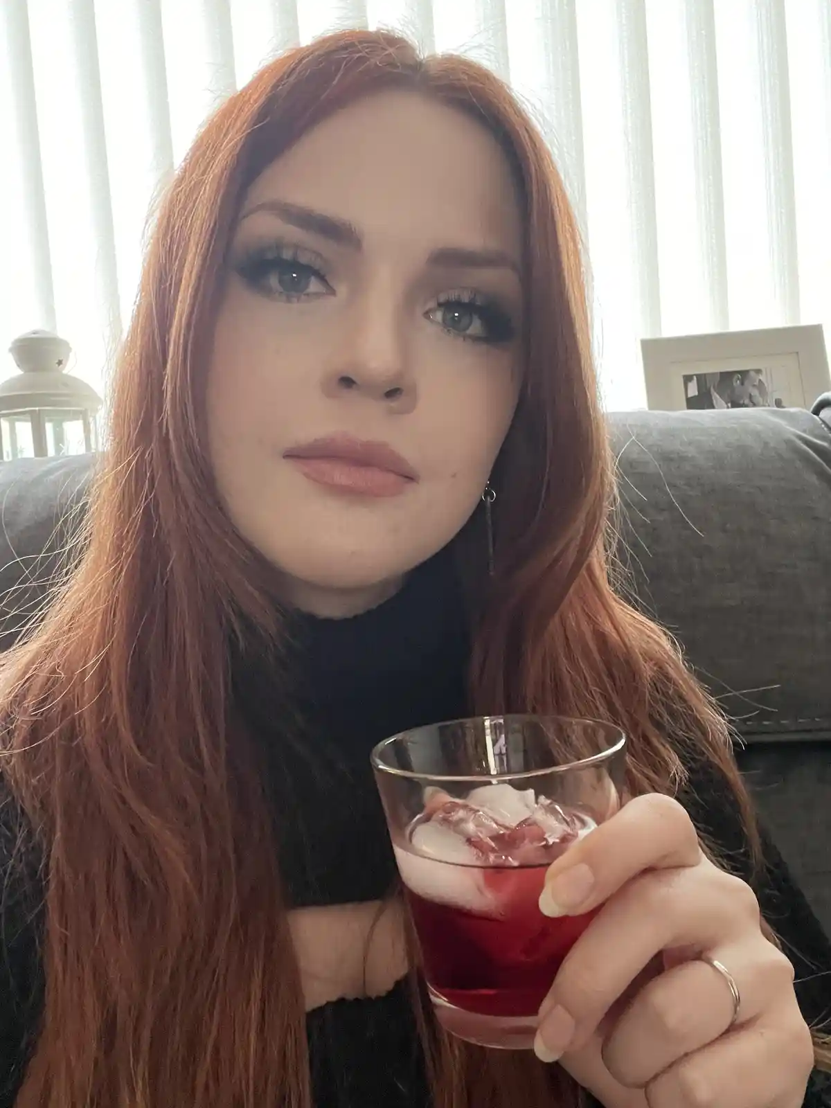 A woman with long red hair and heavy makeup sits on a couch holding a glass of red liquid with ice, looking directly at the…