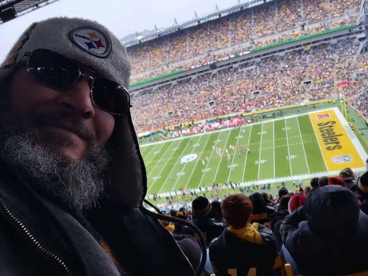 A man with a gray beard, sunglasses, and a Steelers-branded winter hat takes a selfie at a packed Pittsburgh Steelers…