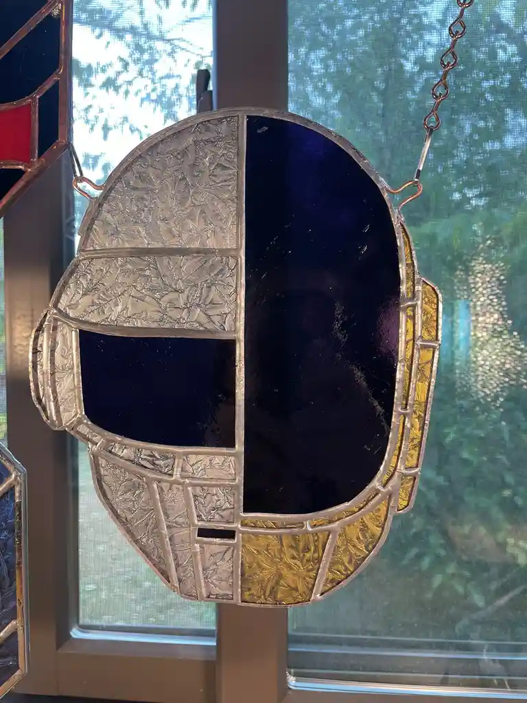A stained glass artwork shaped like a hockey goalie mask hangs by a chain in front of a window with a screen. The mask is…