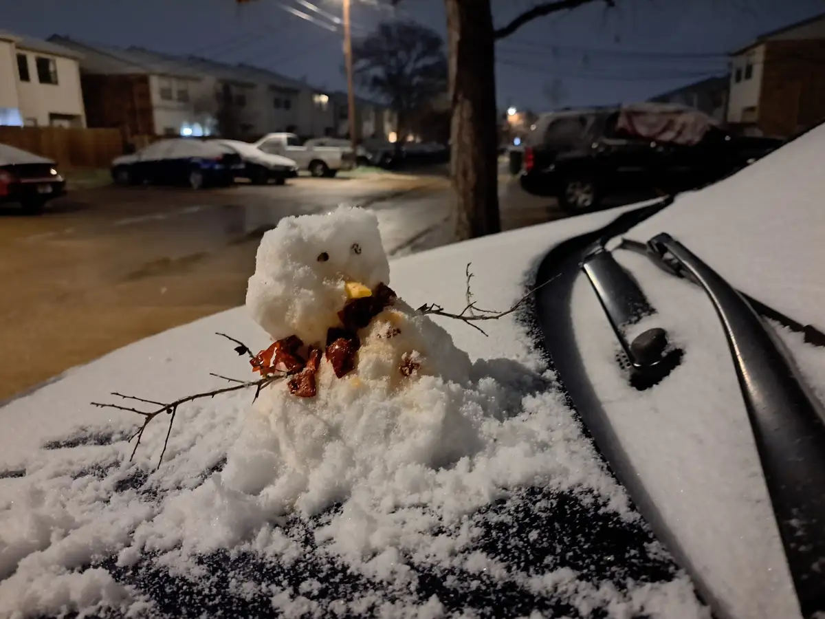 A small, crudely made snowman sits on the snow-covered hood of a car at night, using twigs for arms and what appears to be…