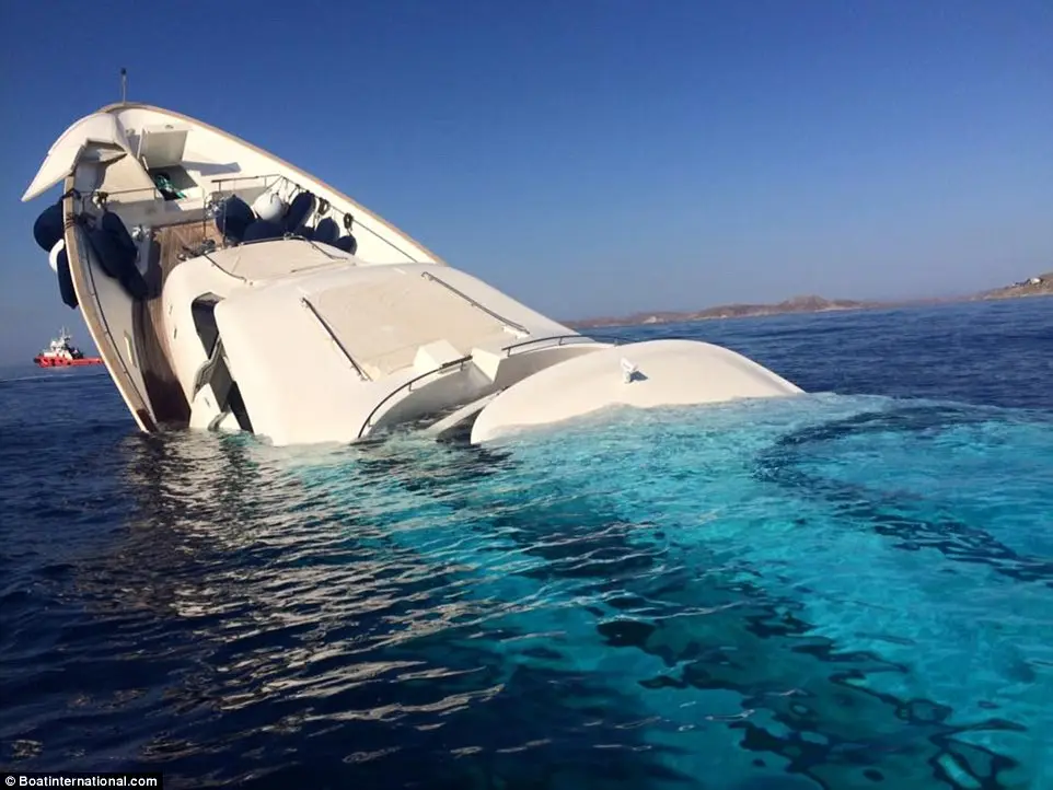 A white luxury yacht is severely tilted, partially submerged in clear blue water under a bright blue sky, with a small red…