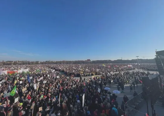 A wide-angle photo shows a massive outdoor crowd of people gathered in a public square under a clear blue sky, many holding…