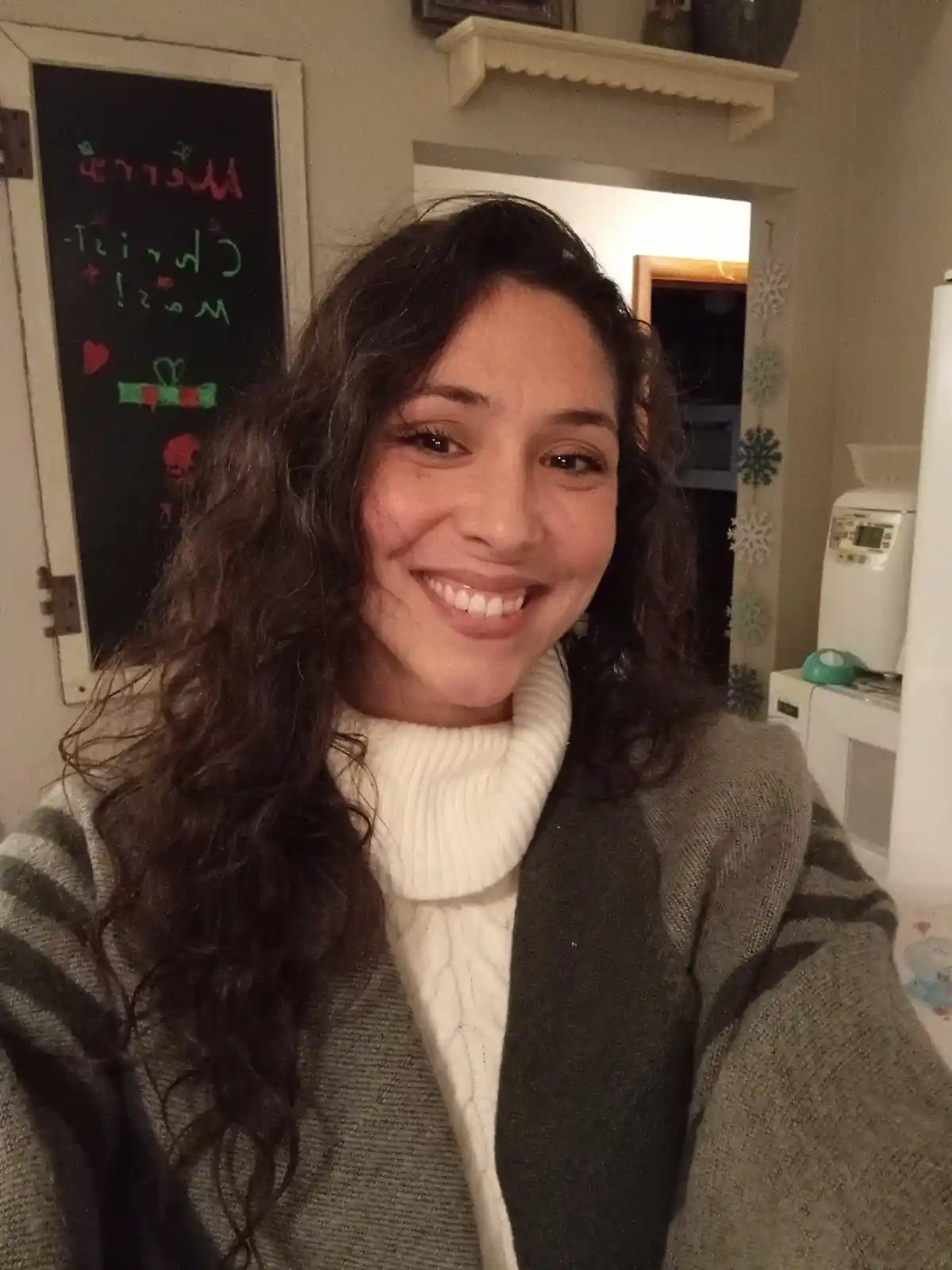 A woman with long dark curly hair smiles at the camera while wearing a white turtleneck and a gray cardigan. Behind her, a…