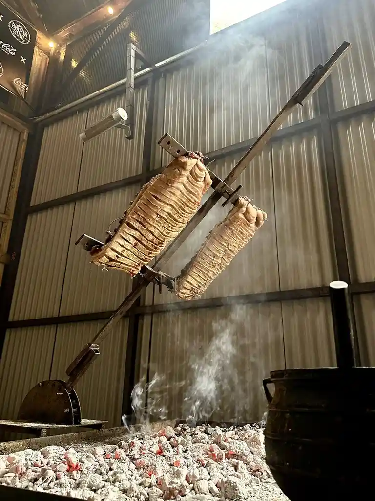 Two large, roasted pork carcasses are suspended on a spit over glowing embers in a rustic, industrial-style smokehouse…