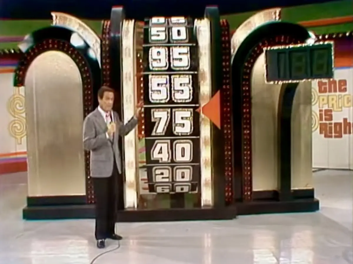 A man in a suit stands beside a large game show pricing wheel displaying numbers 50, 95, 55, 75, 40, 20, and 60. An arrow…