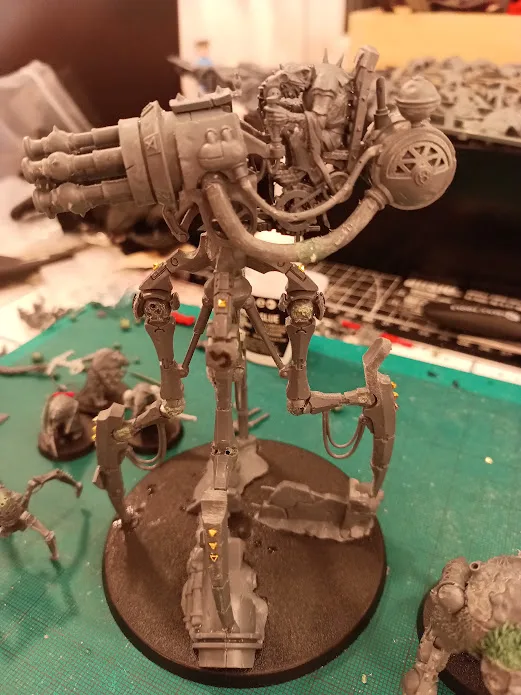 A gray, heavily detailed, multi-limbed robot miniature stands on a circular base, surrounded by other unpainted miniatures…