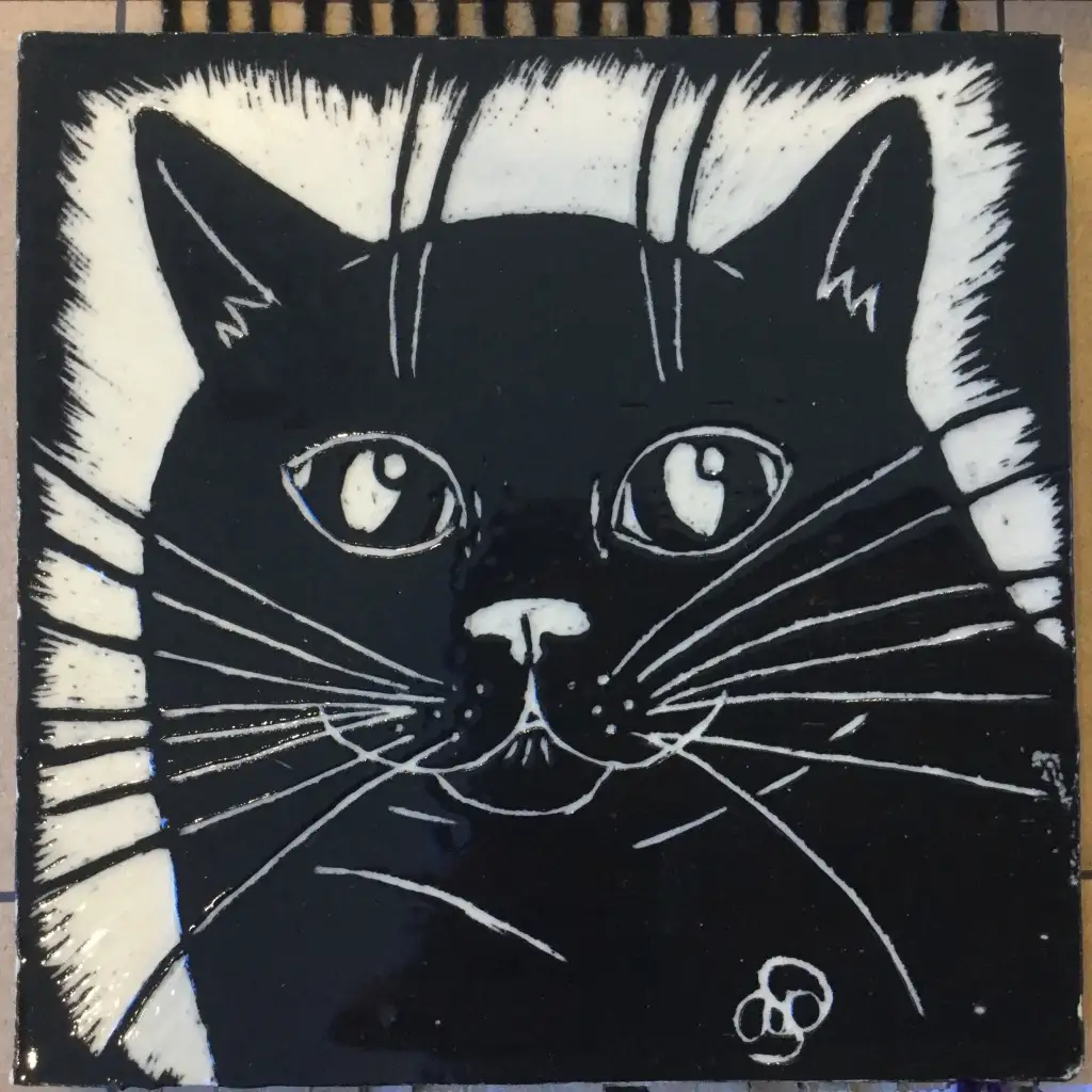 A black-and-white ceramic tile features a stylized illustration of a cat's face with white whiskers, eyes, and fur markings…