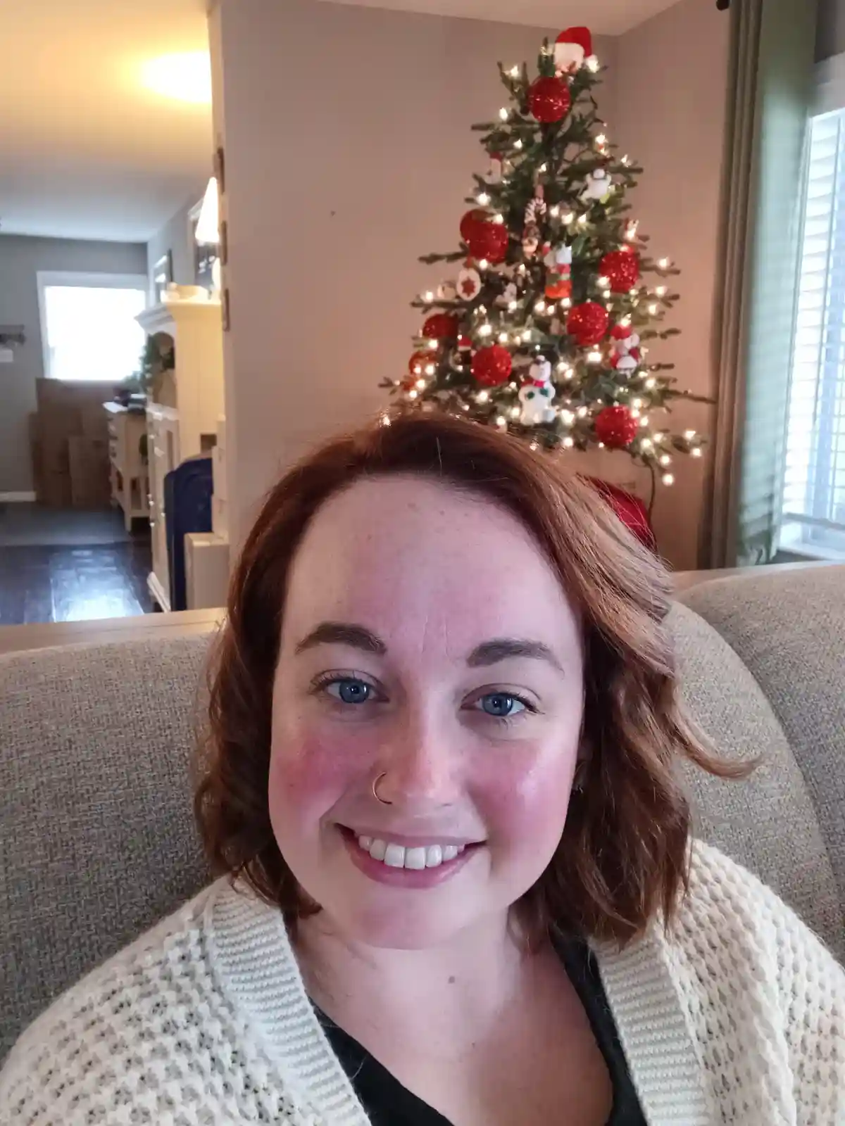 A woman with red hair and blue eyes smiles at the camera while sitting on a couch, with a decorated Christmas tree visible…