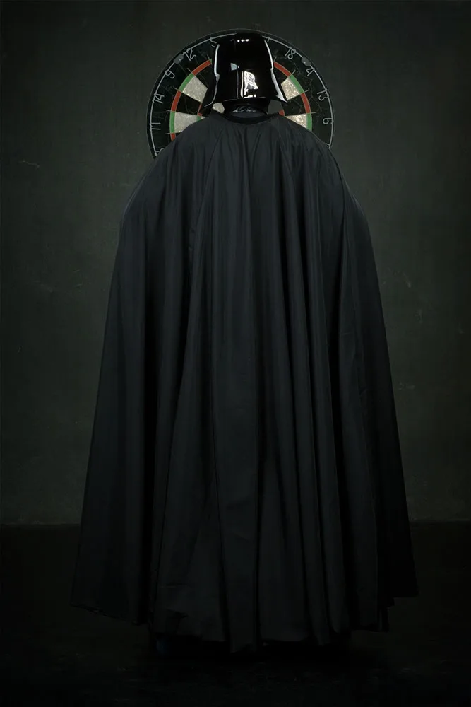 A person in a Darth Vader costume, viewed from behind, stands facing a dartboard mounted on a dark wall. The iconic black…