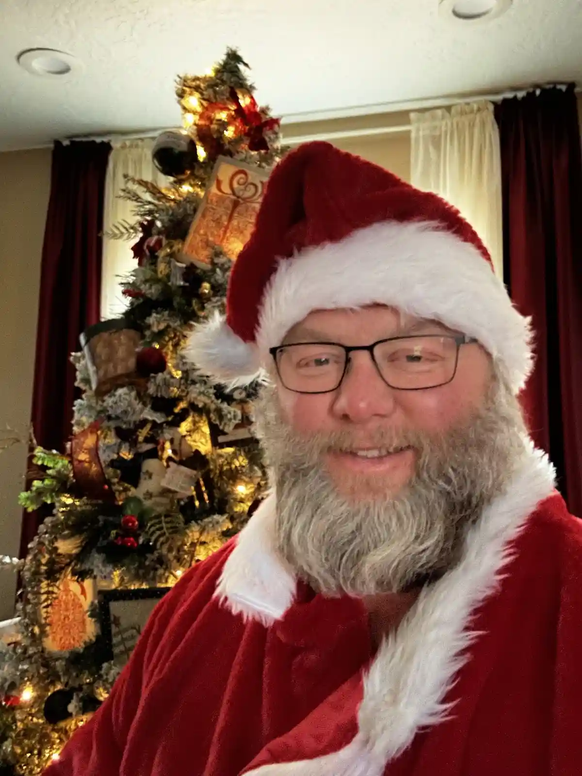A man with glasses and a gray beard wears a red Santa hat and coat with white trim, smiling at the camera. Behind him is a…