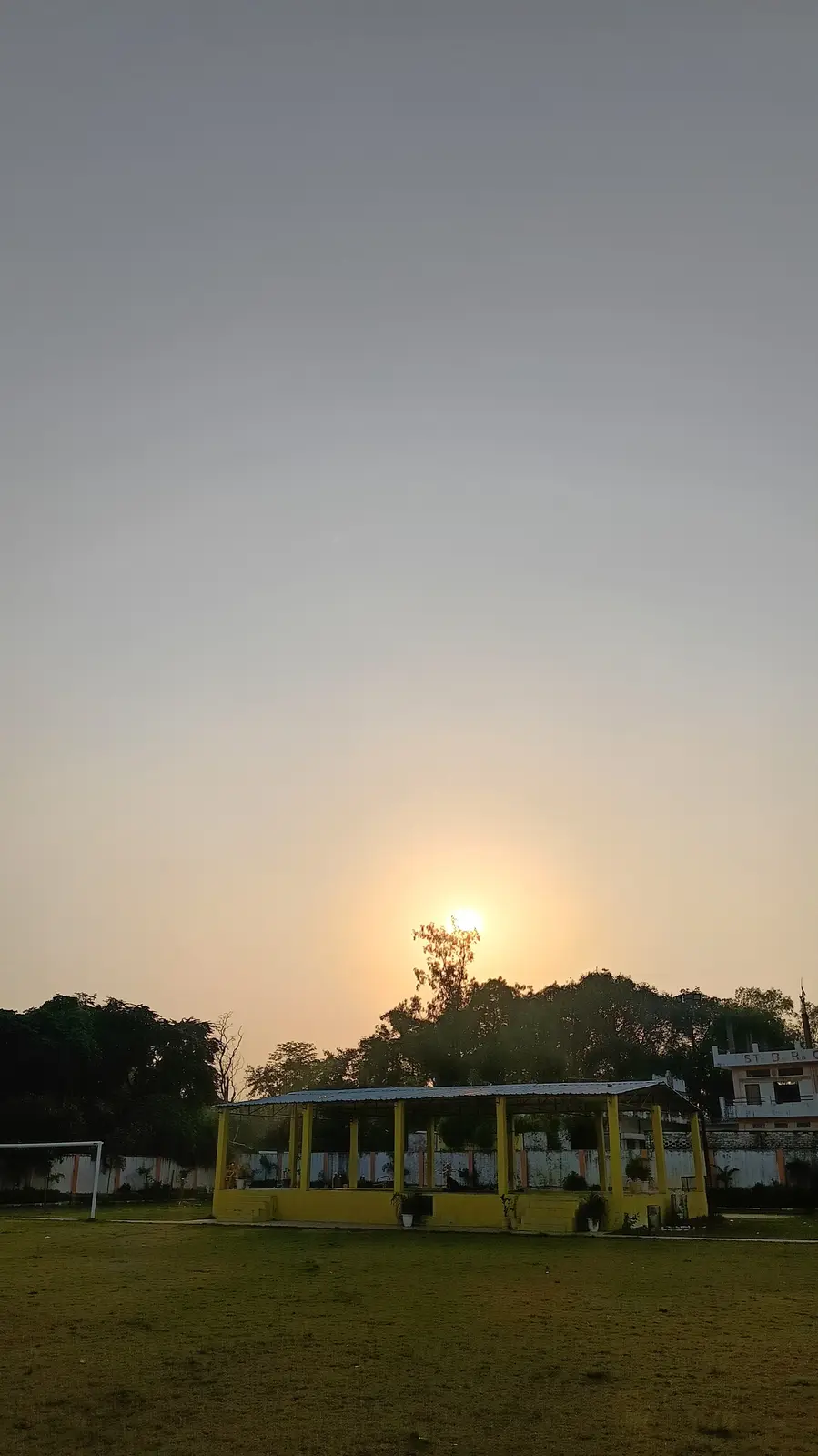A serene outdoor photo captures a sunrise over a grassy school field, featuring a yellow open-sided pavilion and a soccer…