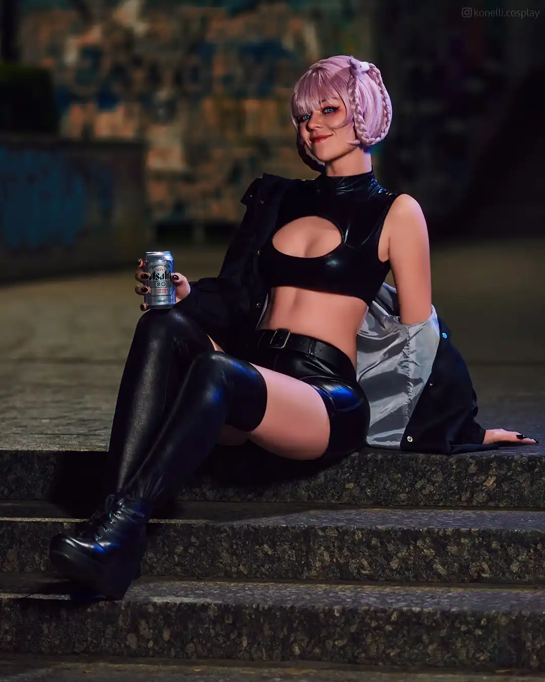 A person in a black leather outfit with pink braided hair sits on stone steps holding a can of Asahi beer, smiling…