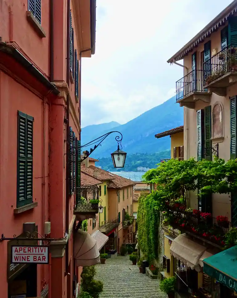 A picturesque, narrow cobblestone street in a European town with colorful buildings, green shutters, balconies with flowers…
