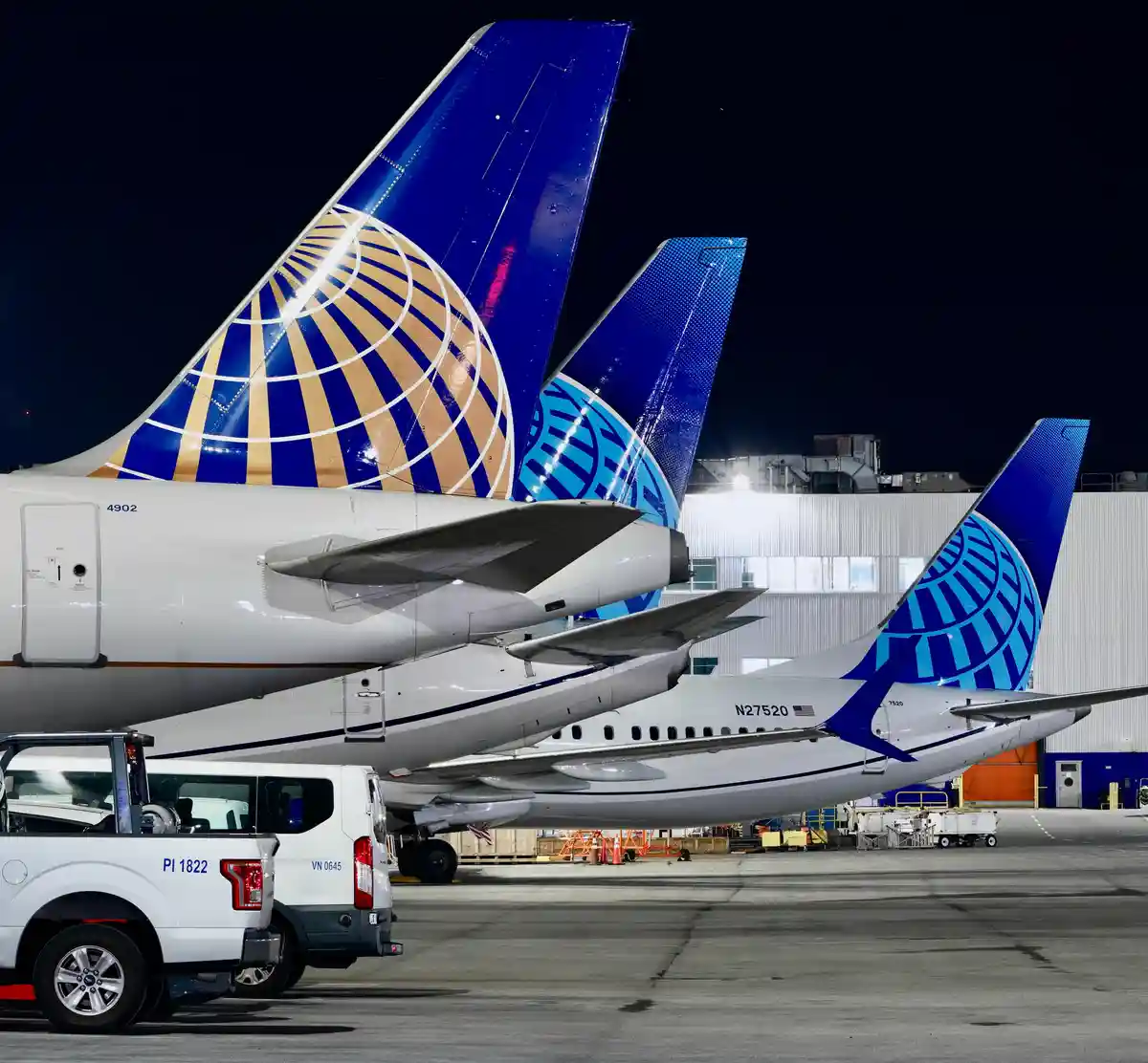 Three United Airlines planes are parked nose-to-tail at an airport at night, their blue and gold tail fins visible against a…