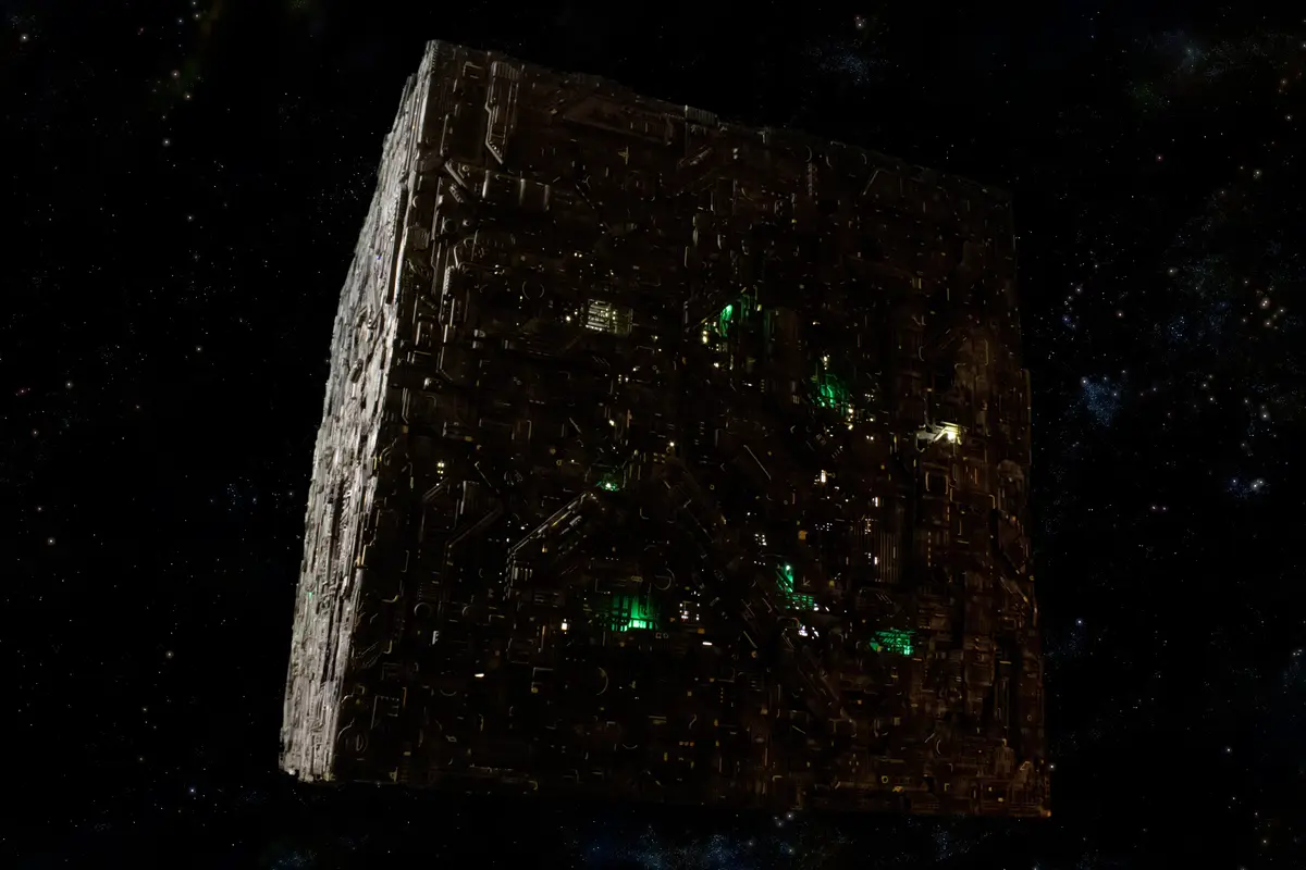 A massive, intricate, dark metallic cube with glowing green and yellow lights floats in a star-filled space background. The…