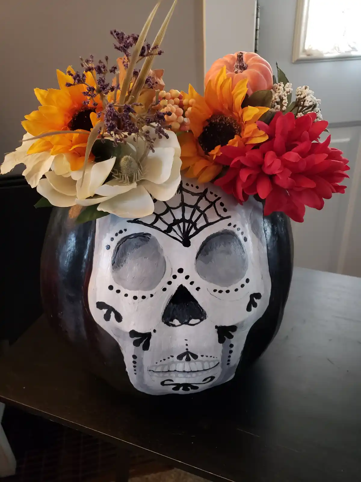 A dark-colored pumpkin is painted with a detailed white and black sugar skull design, featuring decorative patterns around…
