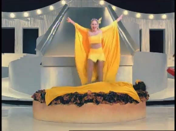 A woman in a bright yellow outfit stands triumphantly with arms raised on a giant, oversized sandwich that has meat and a…