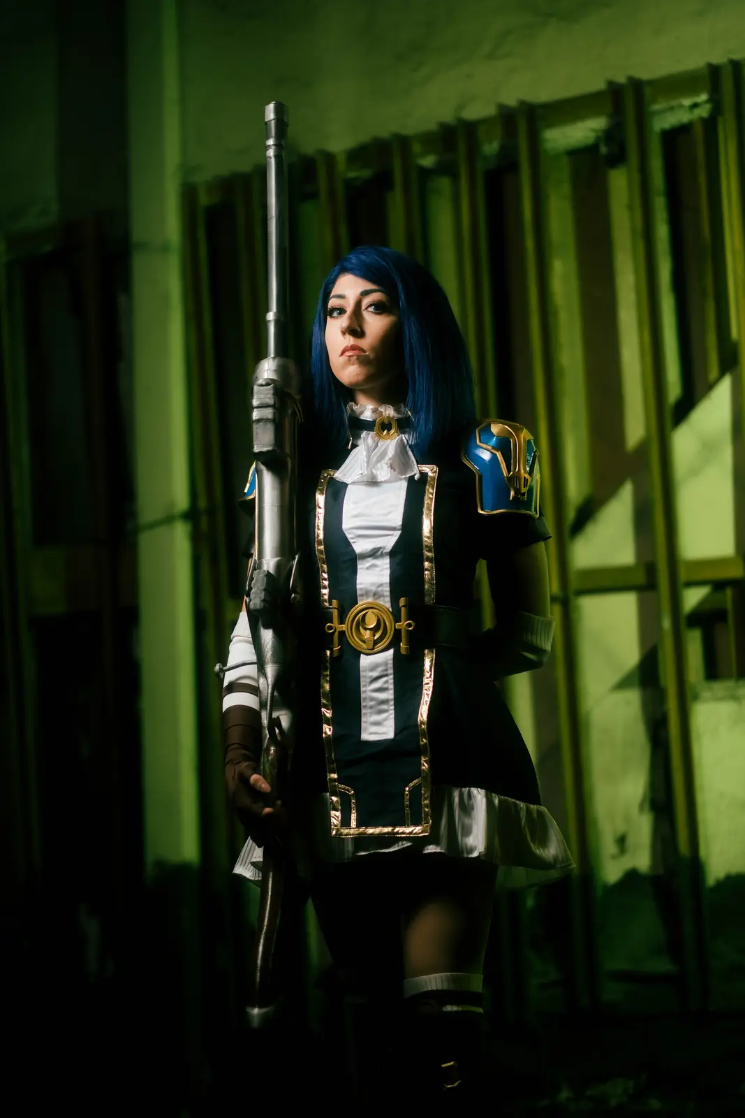 A person in cosplay as a character with blue hair, holding a large rifle, stands in a dimly lit, green-hued room with barred…