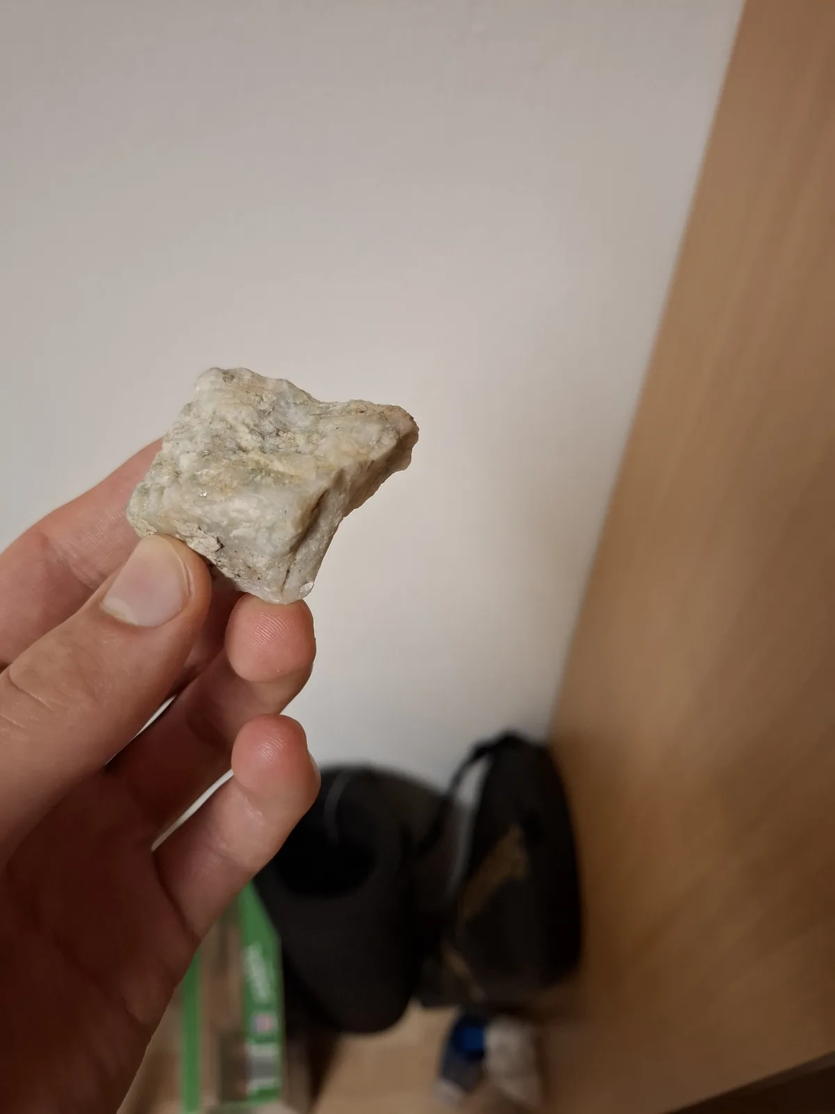 A hand holds up a small, rough, light-gray rock that appears to have a somewhat square or blocky shape. The background is…