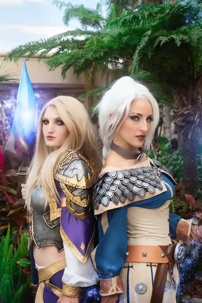 Two women in elaborate fantasy cosplay stand back-to-back outdoors, one holding a glowing blue staff. The woman on the left…