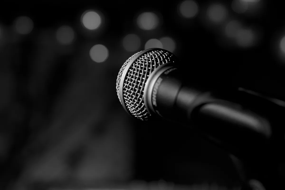 A black and white close-up photograph of a Shure microphone on a stand, with a blurred background of stage lights creating…