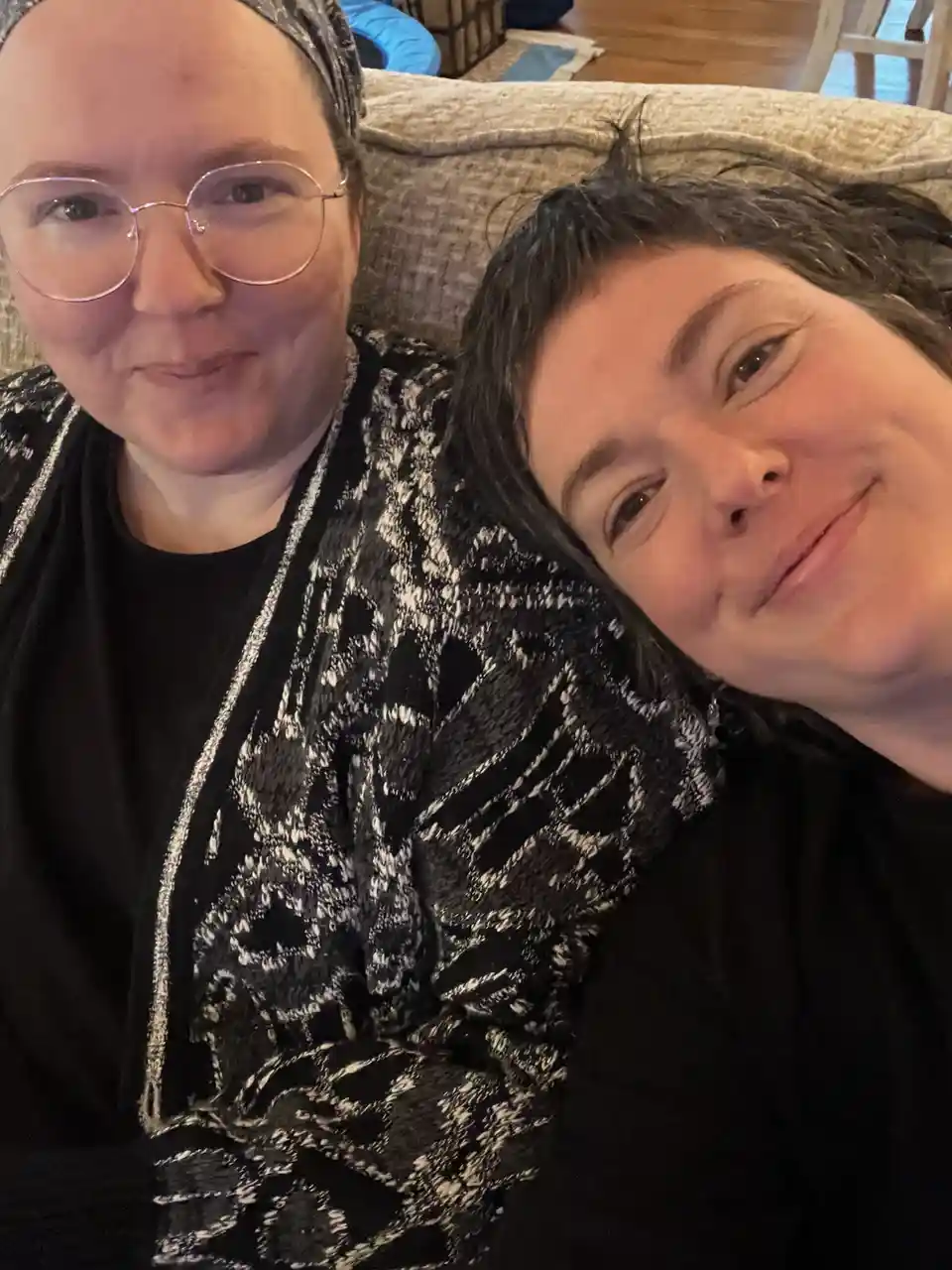 Two women are taking a selfie on a couch; one wears glasses and a black-and-white patterned cardigan, the other has dark…