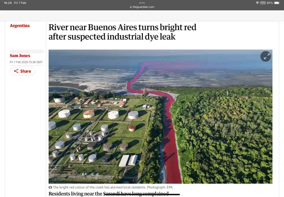 An aerial photo shows a river flowing through a green landscape, its water vividly dyed red, contrasting sharply with the…