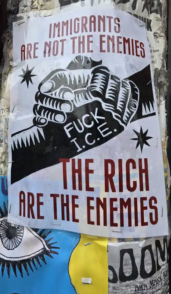 A protest poster with bold red text declares 'IMMIGRANTS ARE NOT THE ENEMIES' at the top and 'THE RICH ARE THE ENEMIES' at…