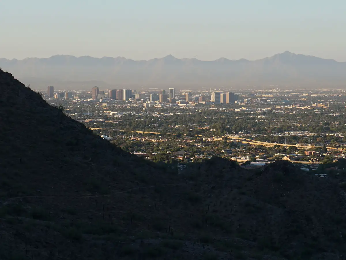A scenic view from a desert hillside overlooking a sprawling city with distant mountains under a hazy sky, overlaid with the…