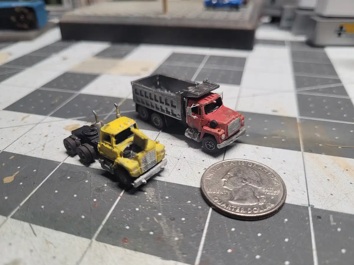 Two miniature model trucks, one yellow and one red, sit on a checkered floor next to a U.S. quarter coin for scale. The…