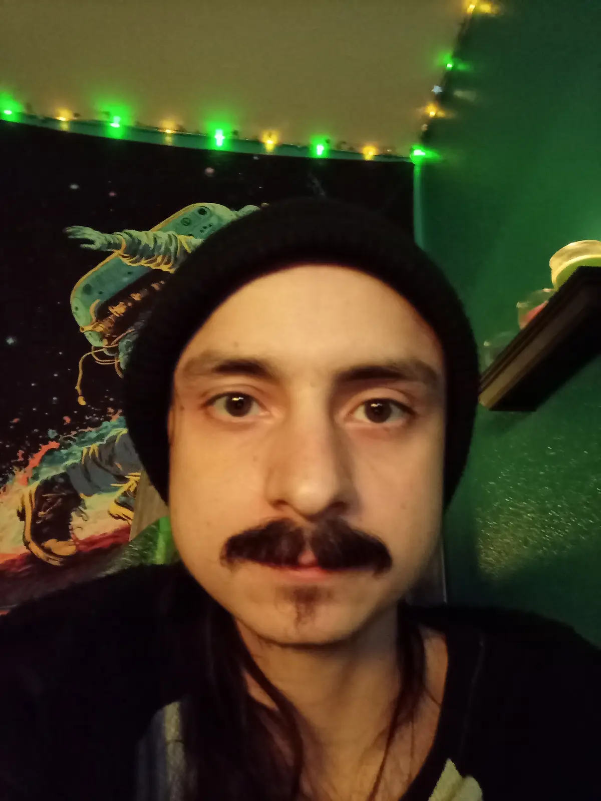 A selfie of a man with a thick, dark mustache and goatee, wearing a black beanie and looking directly at the camera with a…