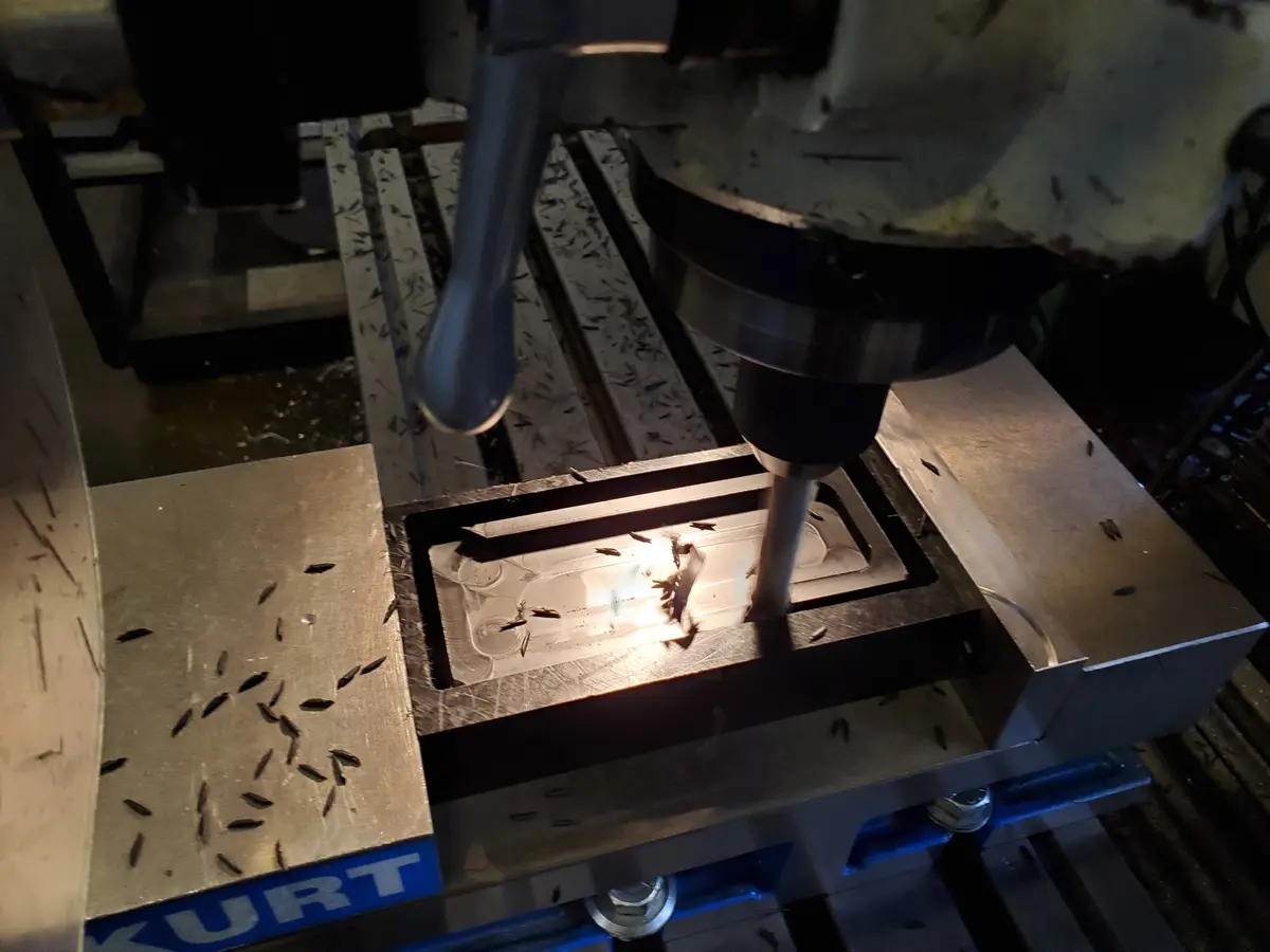 A close-up photo shows a metal drill press actively machining a rectangular metal workpiece, with metal shavings scattered…