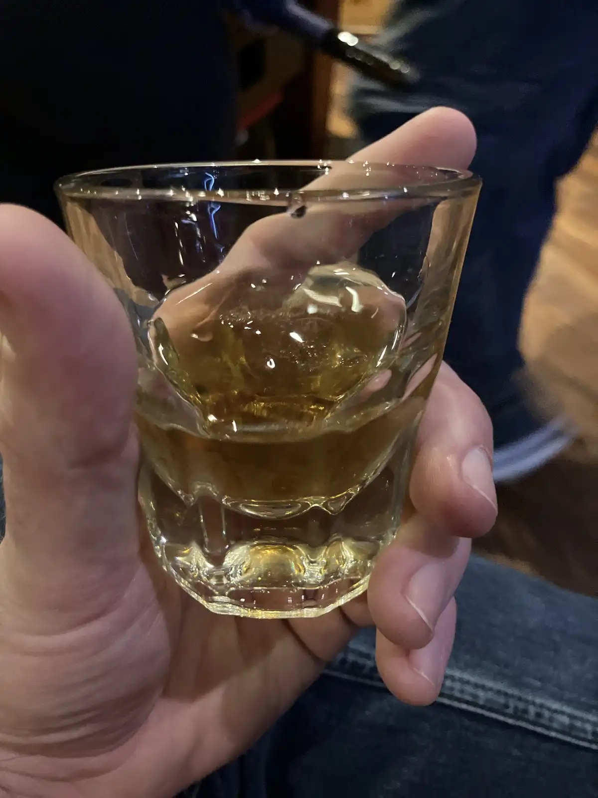 A close-up photo shows a person's hand holding a faceted shot glass containing amber-colored liquid and a large ice cube…