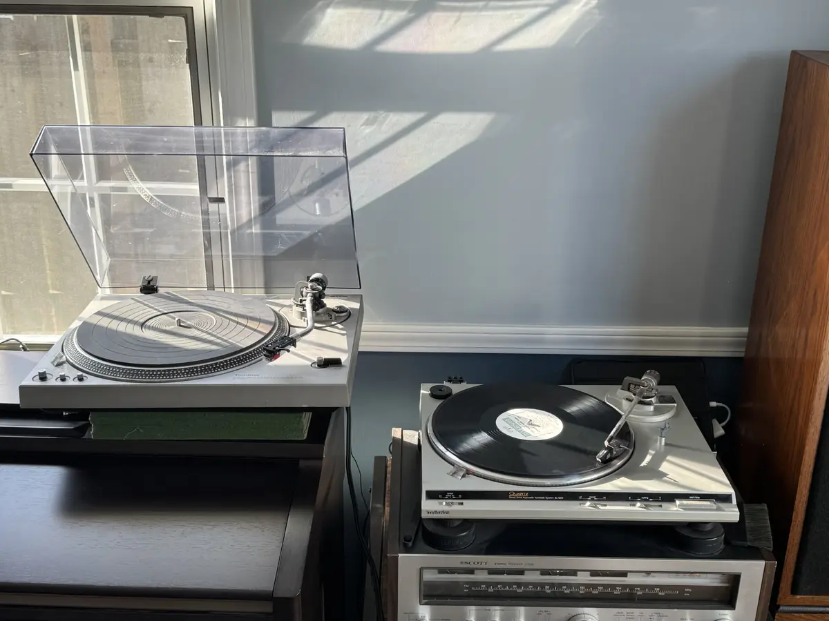 Two vintage Technics turntables sit side-by-side on wooden stands in a room with sunlight casting shadows on the wall behind…
