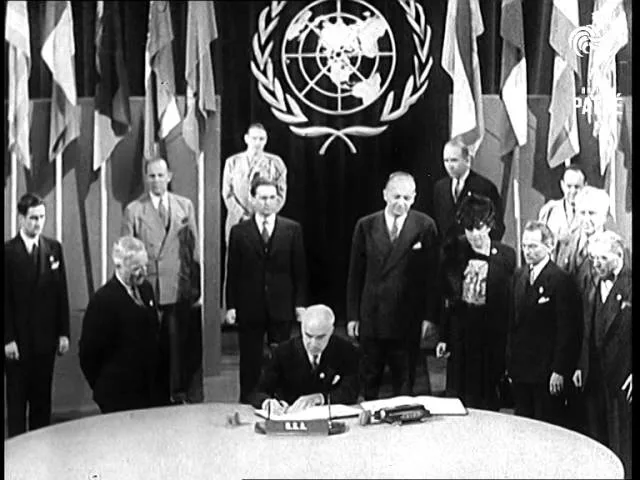 a black-and-white historical photo shows a man in a suit signing a document at a large round table, surrounded by other…