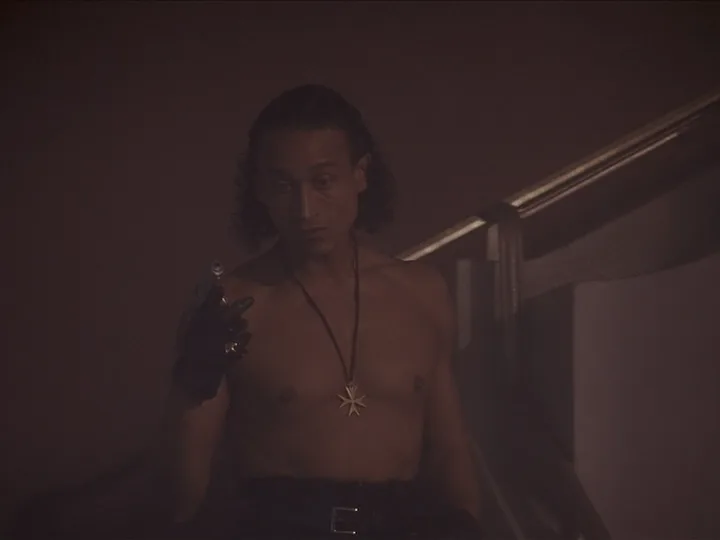 A shirtless man with long dark hair and a black glove holds a small object while wearing a cross pendant; overlaid text…