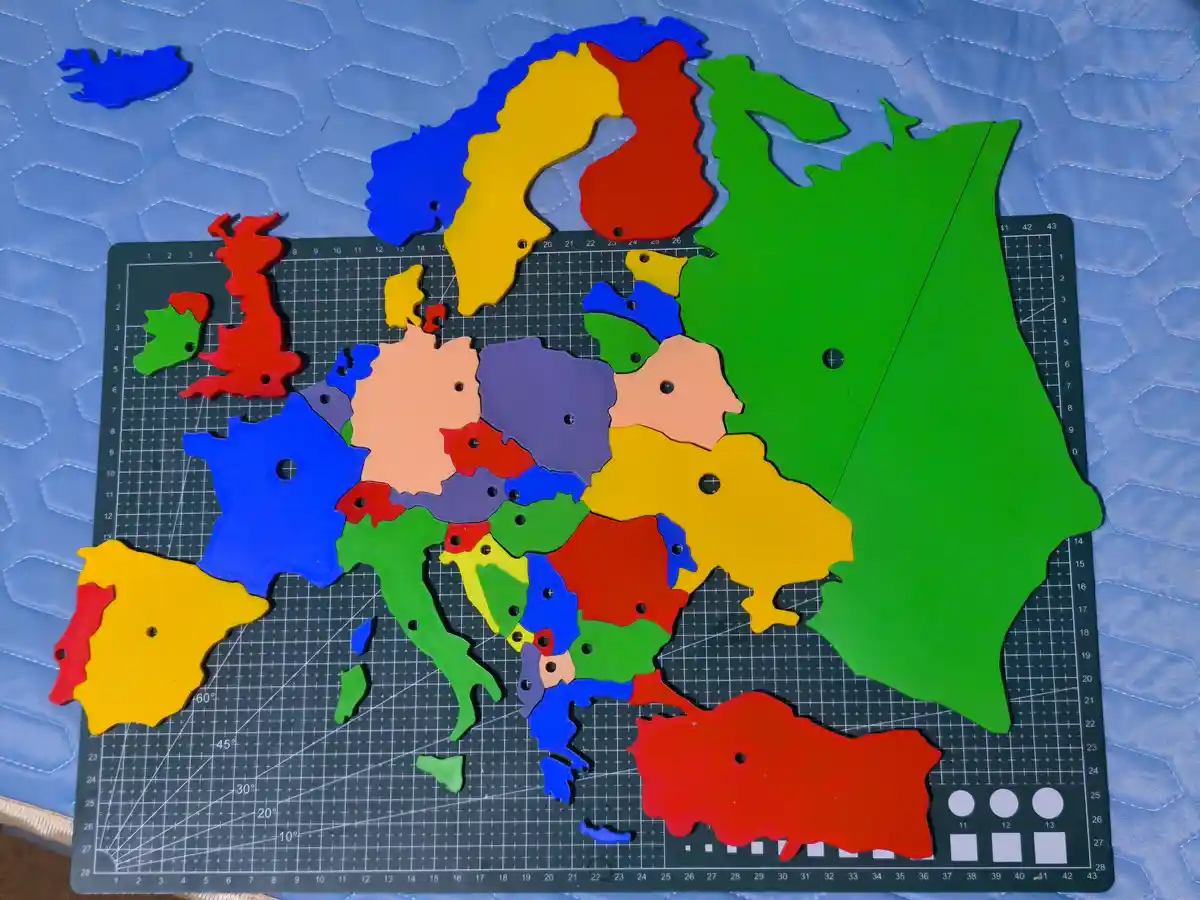 A colorful wooden puzzle map of Europe is laid out on a black gridded cutting mat, with each country piece in a different…