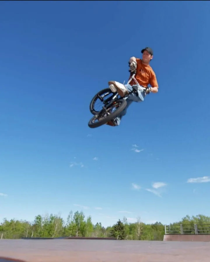 A person in an orange shirt and jeans is airborne on a bicycle against a clear blue sky, with trees and a skatepark visible…