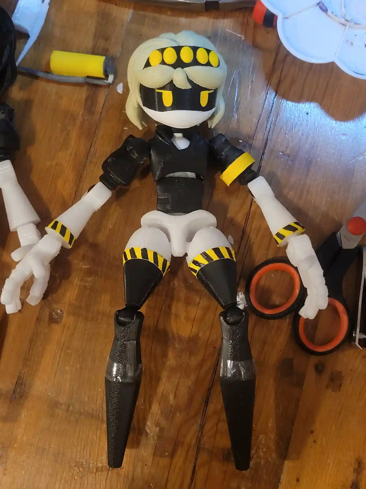 A 3D-printed action figure resembling a character with blonde hair, black and yellow hazard stripes, and a white mask lies…