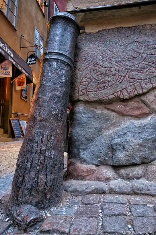 A rusty, weathered cannon is embedded in a cobblestone street, leaning against a stone wall with red Viking-style carvings…